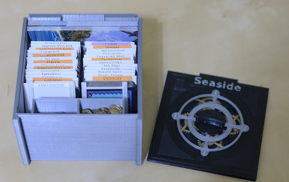 View of an open storage box for Dominion: Seaside with compartments for the cards and other game pieces Black and silver storage box for Dominion: Seaside with the lid off, displaying the cards and other game pieces stored and organized inside